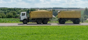 An orange truck with a trailer transports grain along the highway on a beautiful natural landscape. The time of harvesting of grain crops and other sowing and harvesting operations