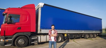 Portrait of professional American truck driver in casual clothing and boots standing in front of truck vehicle with long trailer.